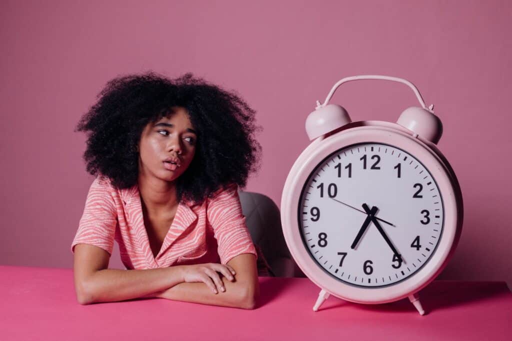 Woman with afro in a pink background staring at a giant alarm clock