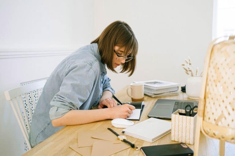 woman writing at a busy desk in front of a laptop