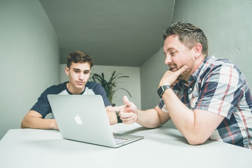 A man pointing at a laptop with a younger guy sitting beside him