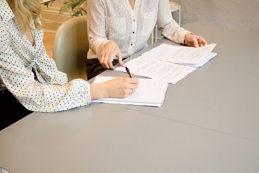 Woman signing on a white paper with another woman sitting beside her